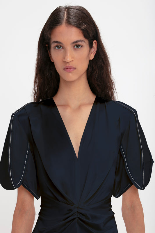 A woman with long brown hair wearing a Victoria Beckham Exclusive Gathered V-Neck Midi Dress In Navy with puffed sleeves, standing against a white background.
