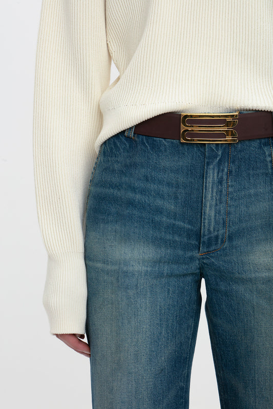 Close-up of a person wearing Victoria Beckham cropped kick jeans in indigrey wash and a cream sweater tucked into a brown belt with a gold buckle.