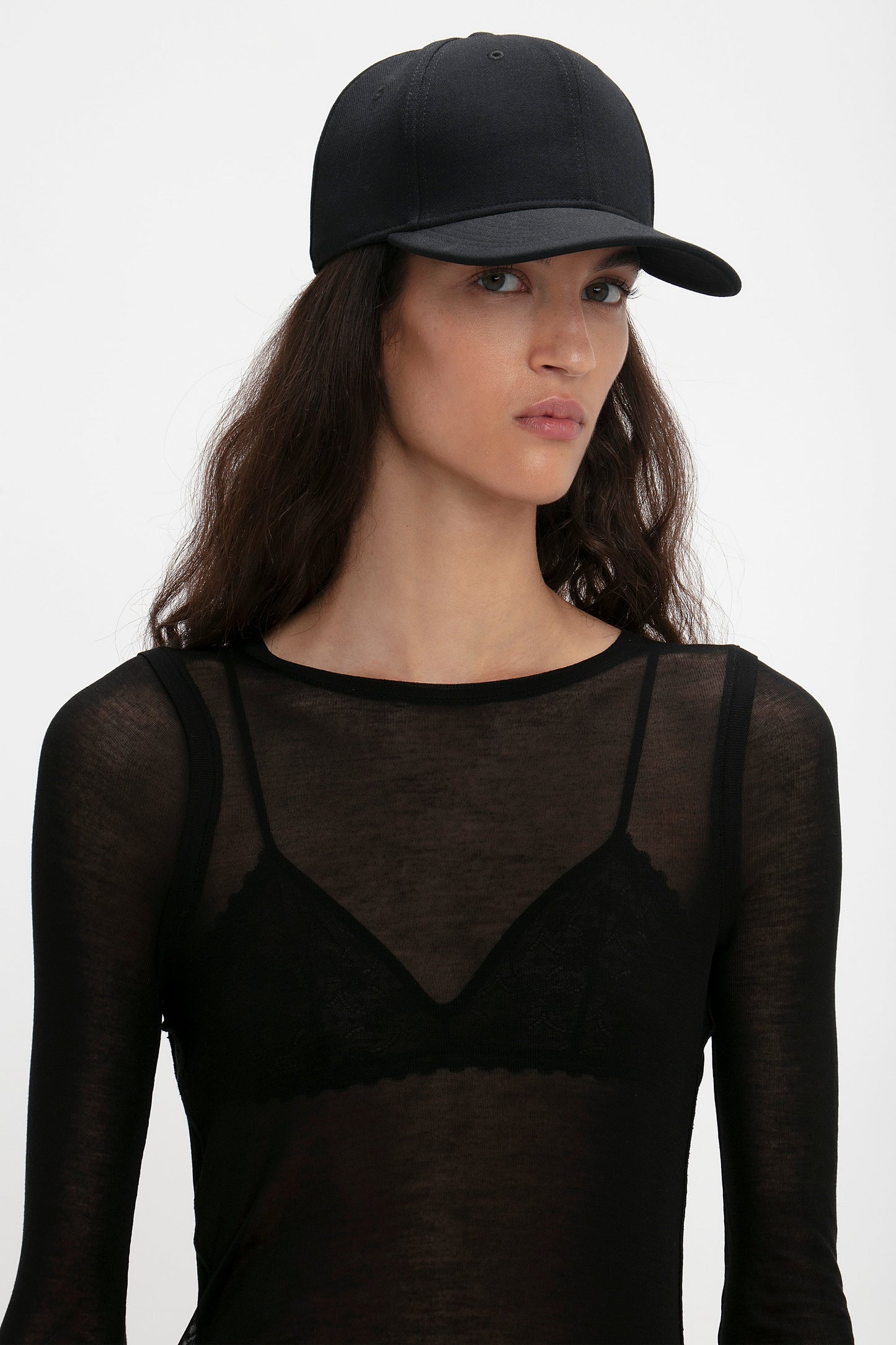 Woman wearing a Victoria Beckham Exclusive Logo Cap in Black and sheer black top with lace details, looking to the side against a white background.