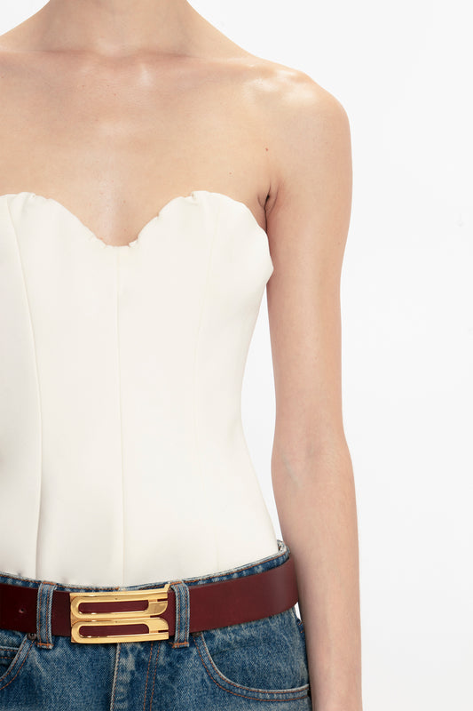 Close-up of a person wearing a Victoria Beckham corset top in antique white with a sweetheart neckline and blue jeans with a brown belt featuring a gold buckle, against a plain background.