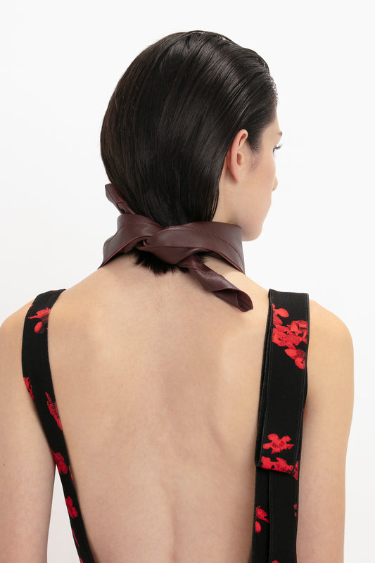 Woman with slicked-back hair wearing a dress with red floral pattern and a Victoria Beckham Foulard In Bordeaux Leather tied at the neck, seen from the back.