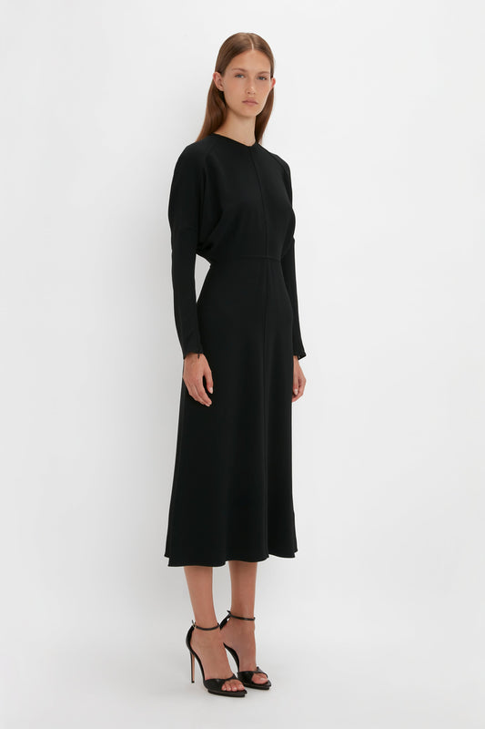 A woman in a simple long-sleeve black Victoria Beckham Dolman midi dress and black high heels standing against a white background.