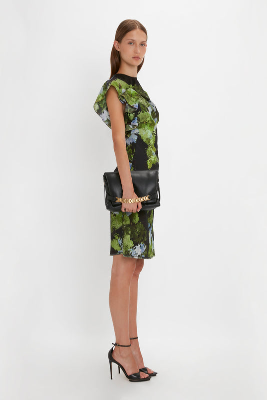 A woman in a green floral dress and black heels carries a Victoria Beckham Puffy Chain Pouch With Strap In Black Leather, posing against a plain white background.