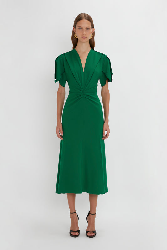 A woman in a Victoria Beckham Gathered V-Neck Midi Dress in Emerald with waist-defining pleat detail and flutter sleeves, standing against a plain white background, wearing black high-heeled sandals.
