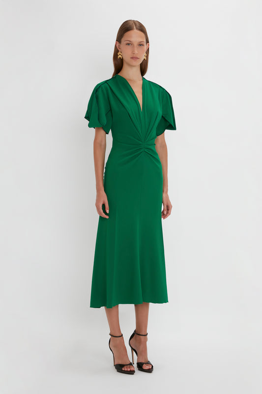 A woman stands in a studio wearing an elegant green Gathered V-Neck Midi Dress in Emerald with puff sleeves and waist-defining pleat detail, paired with black heeled sandals by Victoria Beckham.