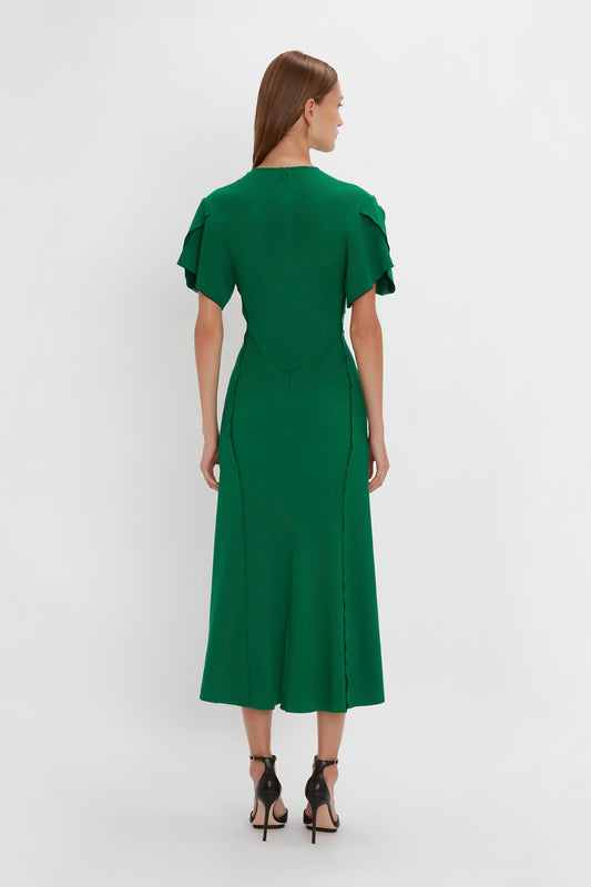 A woman from behind in a Victoria Beckham Gathered V-Neck Midi Dress in Emerald with short ruffle sleeves and black high heels, standing against a white background.