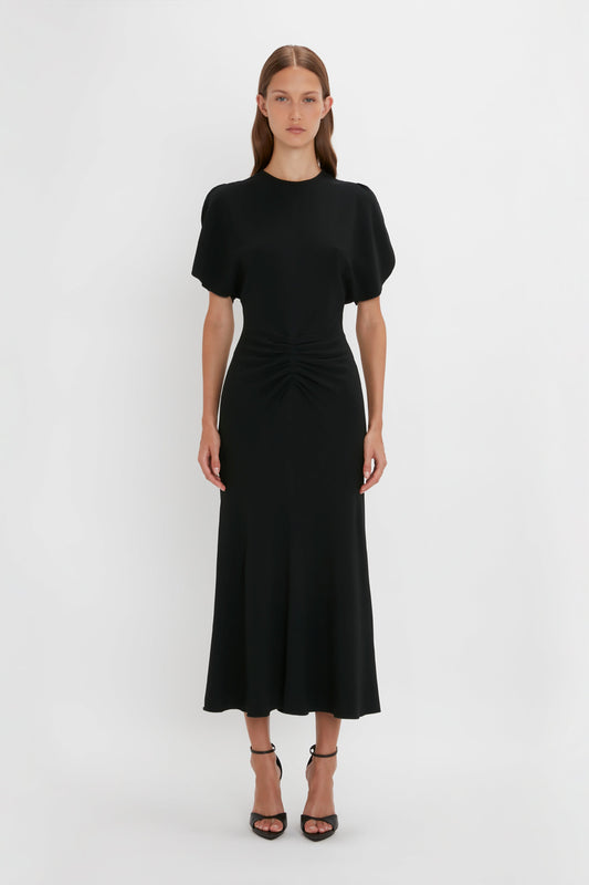 A woman in a Victoria Beckham Gathered Waist Midi Dress In Black with short sleeves and a cinched waist, standing against a white background.