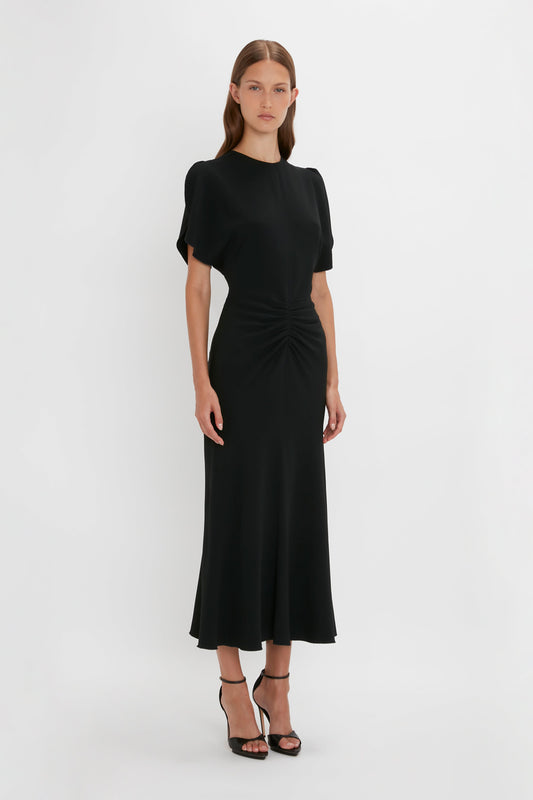 A woman wearing a Victoria Beckham black gathered waist midi dress with short sleeves and a cinched waist, standing against a plain white background. She has on black strappy heels.