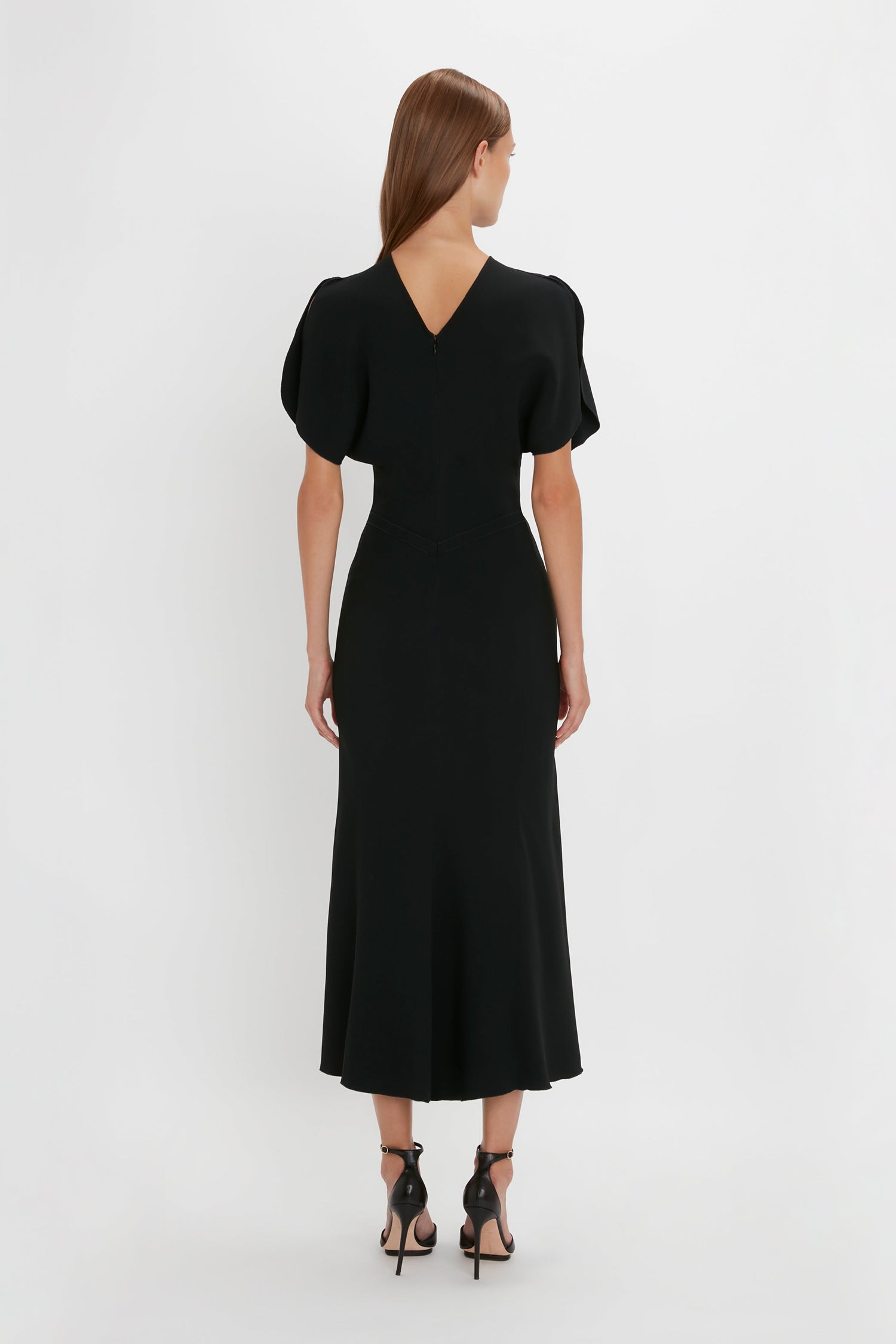 A woman stands facing away, wearing a black Victoria Beckham Gathered Waist Midi Dress with short sleeves and a v-shaped neckline at the back. She is also wearing black pointy-toe stiletto shoes.