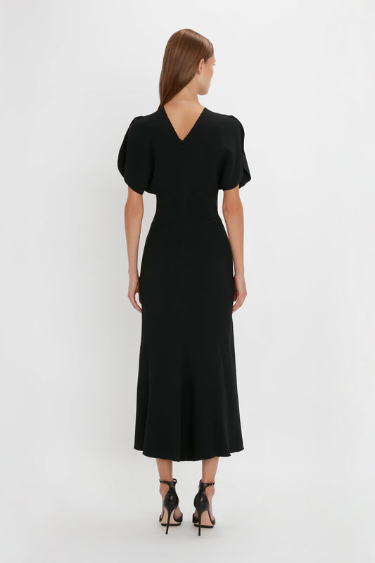 A woman stands facing away, wearing a black Victoria Beckham Gathered Waist Midi Dress with short sleeves and a v-shaped neckline at the back. She is also wearing black pointy-toe stiletto shoes.