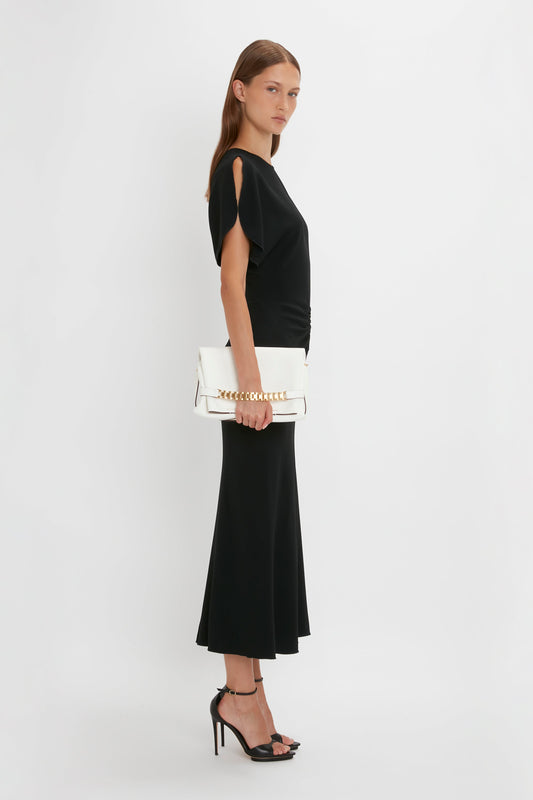 A woman in a Victoria Beckham gathered waist midi dress holding a white handbag stands in profile against a white background.