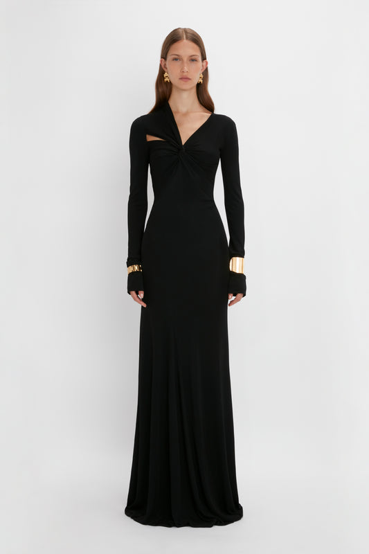 A woman wearing a long black evening dress with a gathered chest design and long sleeves, accessorized with Victoria Beckham contemporary gold-plated brass earrings and bracelets, standing against a white background.