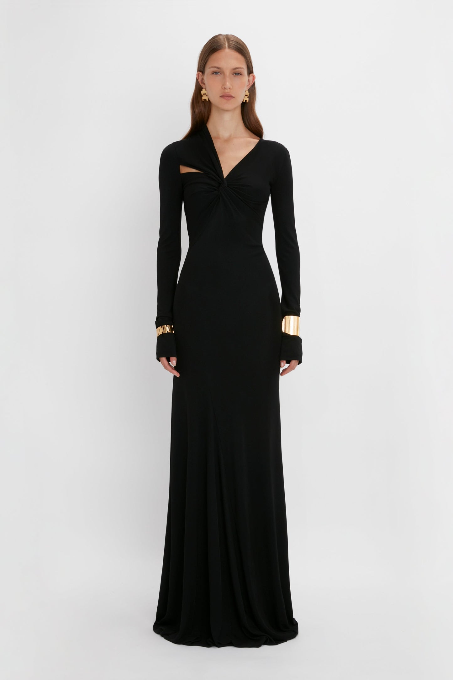 A woman in a black long-sleeve Victoria Beckham floor-length gown with a twisted detail at the chest, accessorized with gold earrings and bracelets, standing against a white background.