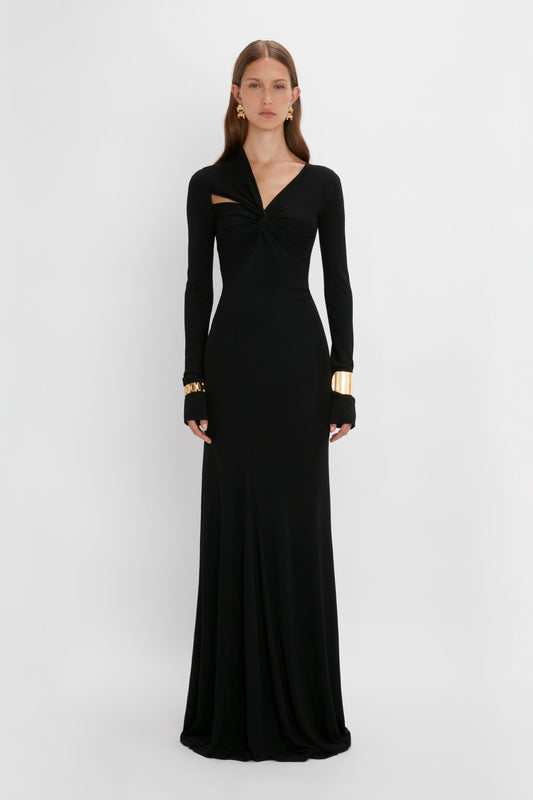 A woman in a black long-sleeve Victoria Beckham floor-length gown with a twisted detail at the chest, accessorized with gold earrings and bracelets, standing against a white background.