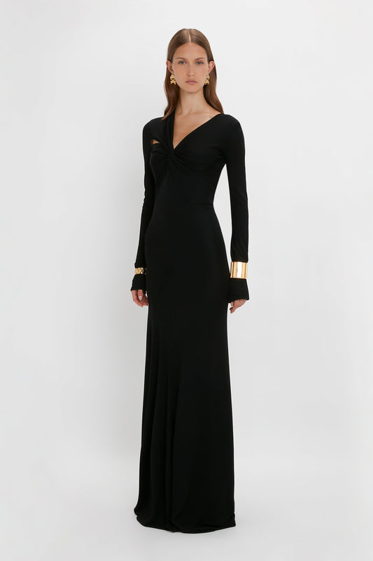 A woman in a sleek, long black "Tie Detail Floor-Length Dress" by Victoria Beckham with long sleeves and gold cuff accessories stands against a white background.