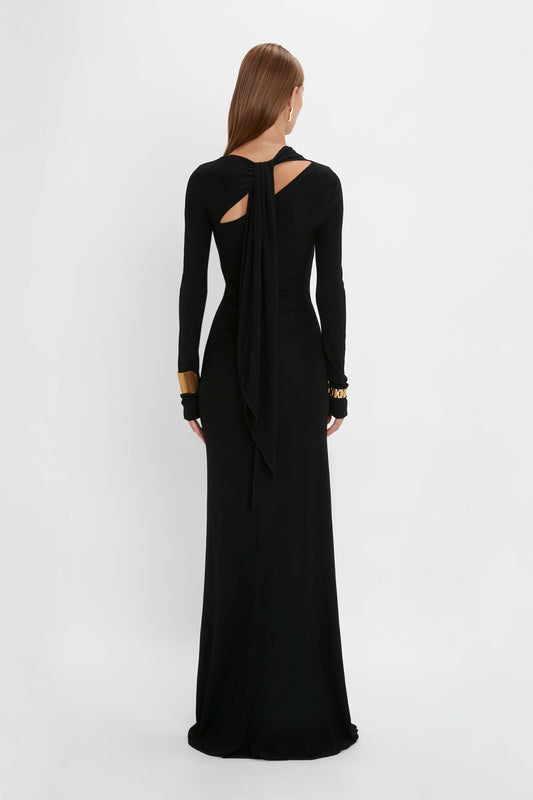 A woman in a Victoria Beckham tie detail floor-length dress in black with a draped, open back design, standing against a white background, viewed from behind.