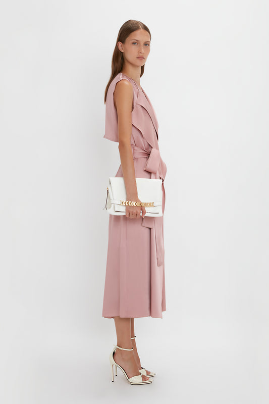 A woman in a pink sleeveless jumpsuit holding a white Victoria Beckham Chain Pouch with Strap In White Leather shoulder bag, standing against a white background.