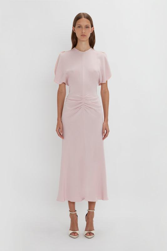 A woman in an elegant soft pink Victoria Beckham midi dress with tulip sleeves and a twisted detail at the waist, standing against a plain white background.