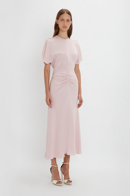 A woman in a Victoria Beckham Gathered Waist Midi Dress In Blush with a twisted front detail and short tulip sleeves, standing against a white background.