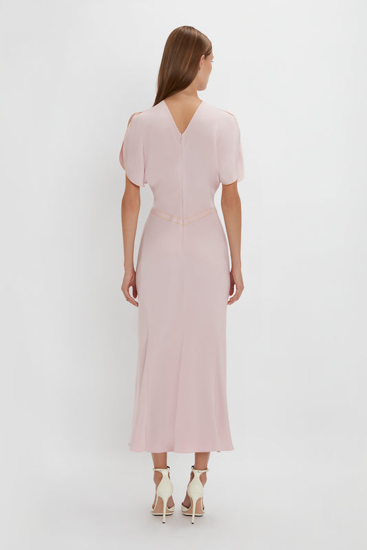 A woman seen from the back wearing an elegant Victoria Beckham Gathered Waist Midi Dress In Blush with tulip sleeves and white high heels against a plain background.