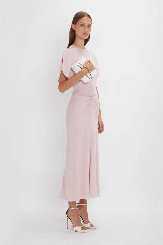 A woman in a Victoria Beckham Gathered Waist Midi Dress In Blush with a V-neck at the back and tulip sleeves, paired with white heels, holding a clutch, posing against a plain white background.