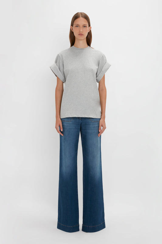 Woman standing upright wearing a Victoria Beckham Asymmetric Relaxed Fit T-Shirt in Grey Marl with wide sleeves and blue bell-bottom jeans against a plain white background.