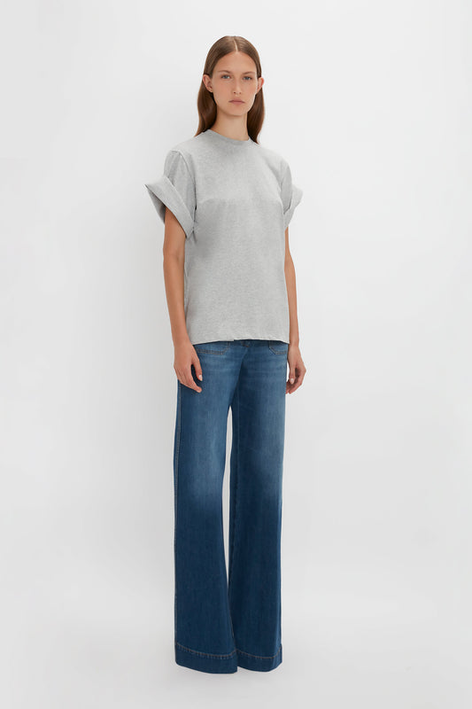 Woman standing in a plain white studio wearing a Victoria Beckham asymmetric relaxed fit T-shirt in grey marl made of organic cotton with voluminous sleeves and blue flared jeans.