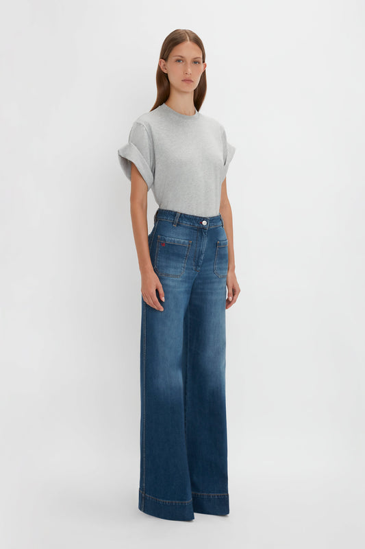 A woman in a Victoria Beckham asymmetric relaxed fit T-shirt in grey marl and high-waisted flared blue jeans standing against a white background.