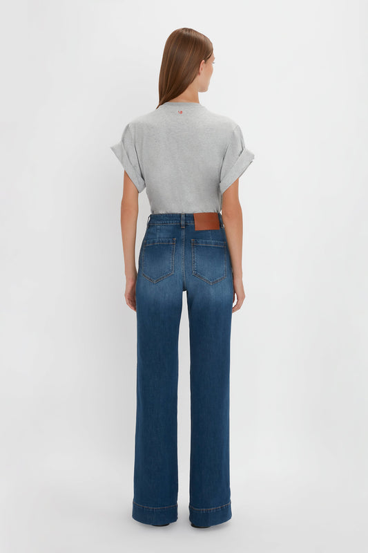 A woman stands facing away from the camera, wearing a gray t-shirt and blue Victoria Beckham Alina jeans in Dark Vintage Wash with a brown belt.
