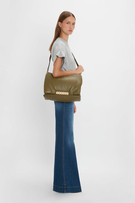 A woman in a gray top and Victoria Beckham Alina Jean in Dark Vintage Wash holds a large olive green shoulder bag and looks over her shoulder in a bright, minimalist setting.