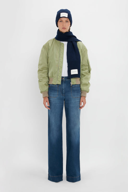 A person in a blue beanie, green bomber jacket with an Exclusive Logo Patch Scarf In Navy from Victoria Beckham, and denim jeans standing against a white background.