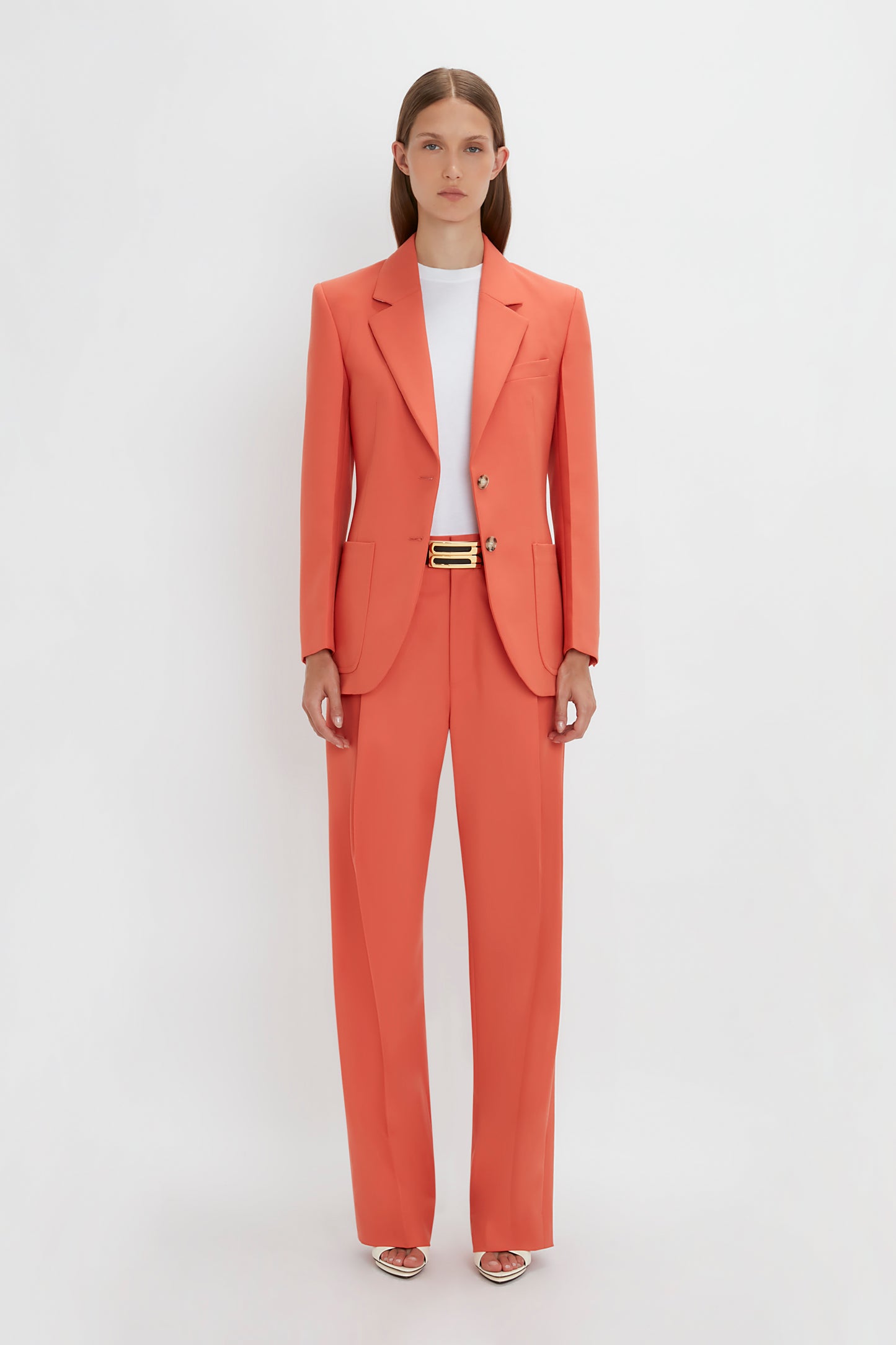 A woman in a bright Patch Pocket Jacket in Papaya by Victoria Beckham with a single-breasted jacket and a black belt, standing against a plain white background.