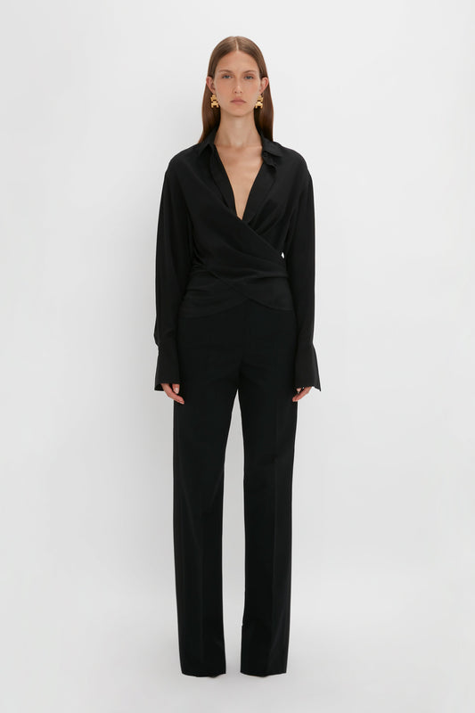 A woman in a Victoria Beckham Wrap Front Blouse In Black and trousers stands against a white background, looking directly at the camera.