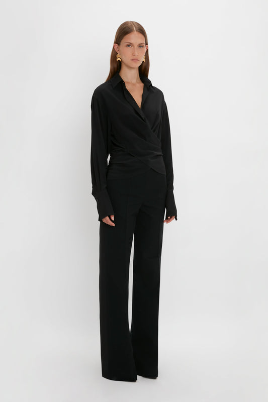 A woman in a Victoria Beckham black silk wrap front blouse and wide-leg trousers stands against a white background.