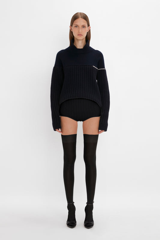 A woman in a black turtleneck sweater dress and Victoria Beckham Exclusive Over The Knee Socks In Black stands against a white background.