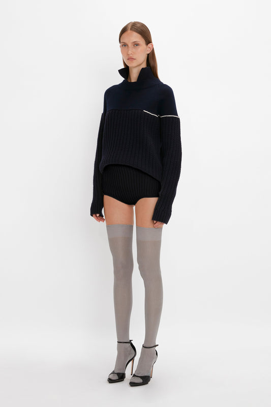 A woman in a dark navy oversized turtleneck sweater paired with Victoria Beckham's Exclusive Over The Knee Socks In Grey and black high-heeled shoes, standing against a white background.