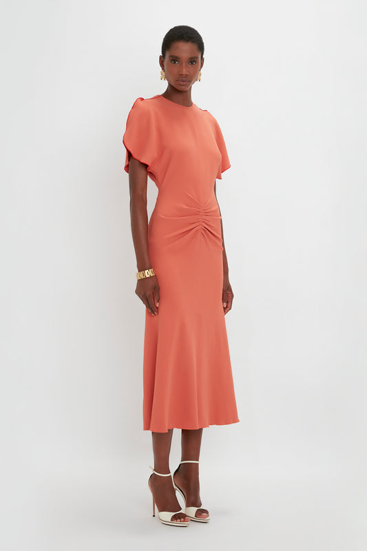 A black woman in a Victoria Beckham Gathered Waist Midi Dress In Papaya and white pointy toe stiletto sandals, standing against a white background.