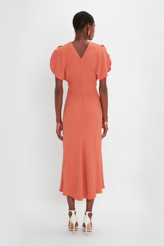 Woman from behind wearing a Victoria Beckham papaya-colored Gathered Waist Midi Dress with puffed sleeves and white pointy toe stiletto sandals, standing against a white background.