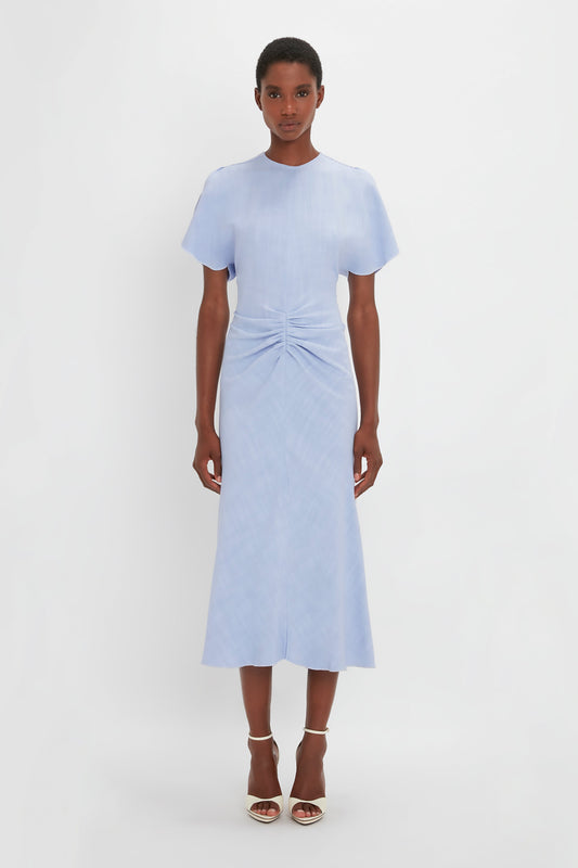 A black woman modeling a Victoria Beckham pale blue Gathered Waist Midi Dress In Frost with short sleeves and a twisted front detail, paired with white pointy toe stiletto sandals, against a plain white background.