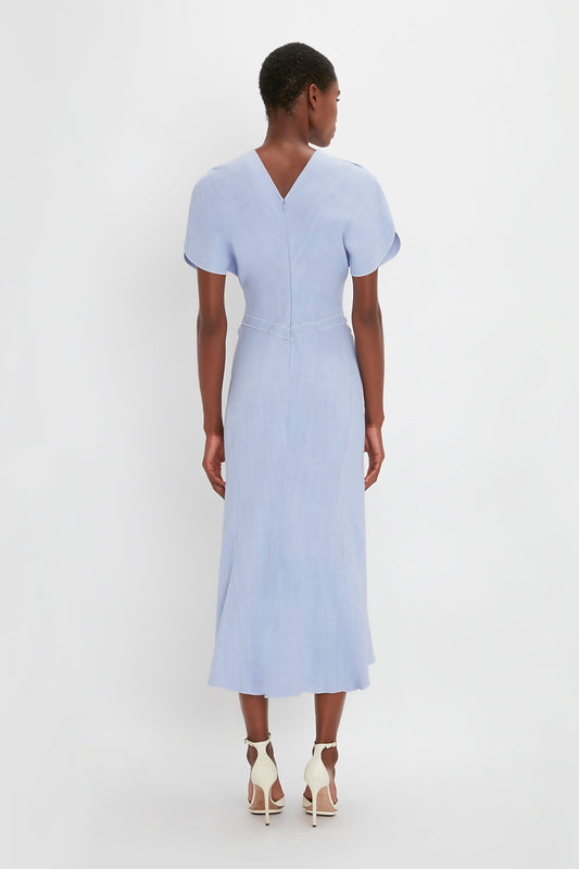 A woman viewed from the back wearing a Victoria Beckham light blue Gathered Waist Midi Dress In Frost with short sleeves and white pointy toe stiletto sandals, standing against a plain white background.