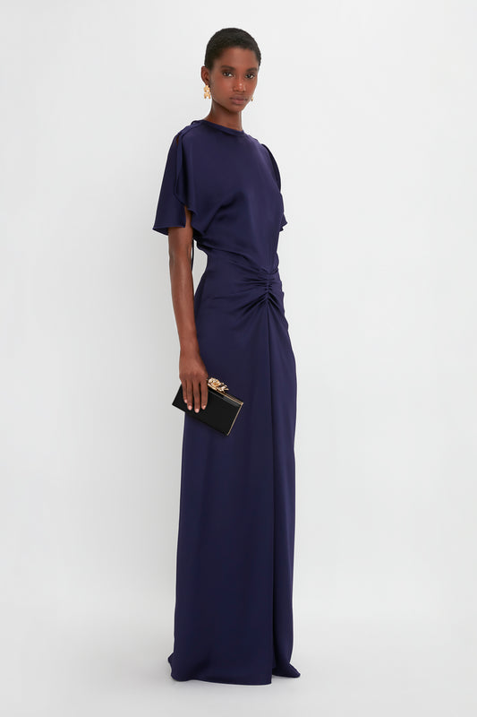 A woman in a dark blue evening gown with a twisted waist detail and short sleeves, holding a Victoria Beckham Frame Flower Minaudiere in Black clutch with a brass frame, standing against a plain white background.