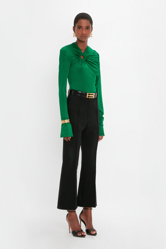 A woman stands modeling a green blouse with a twist detail and black trousers, accessorized with a Victoria Beckham Exclusive Jumbo Chain Bracelet in Brushed Gold and black heels, against a white background.