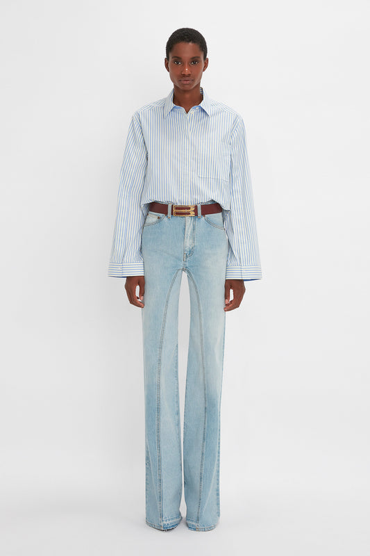 A young man wearing a striped light blue and white button detail cropped shirt and Victoria Beckham's Bianca Jean In Light Blue Denim with a brown belt, stands against a white background.