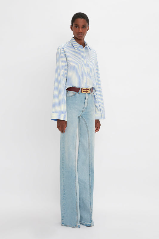 A woman stands facing the camera, wearing a striped blue and white button detail cropped shirt, light blue wide-leg Victoria Beckham Bianca jeans, and a brown belt.