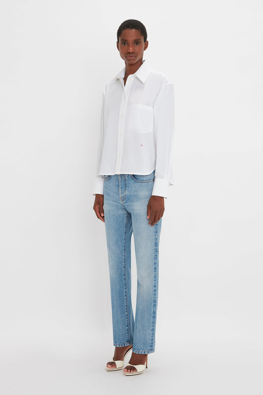 A woman in a classic white Cropped Long Sleeve Shirt In White by Victoria Beckham monogram and blue jeans stands facing the camera against a plain white background.