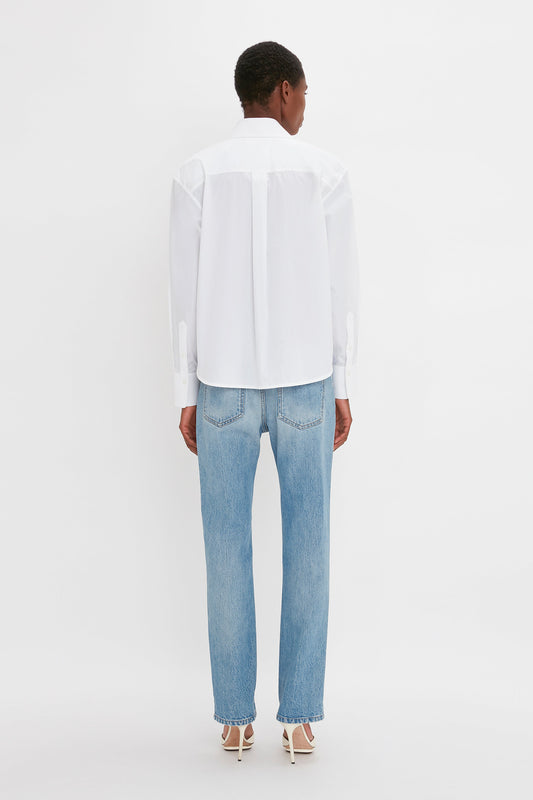 A woman from behind, wearing a Victoria Beckham cropped long sleeve shirt in white and blue jeans, stands against a plain white background.