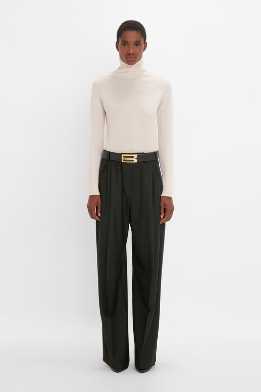 A person wearing a beige Polo Neck Victoria Beckham sweater and black trousers with a gold belt, standing against a plain white background.