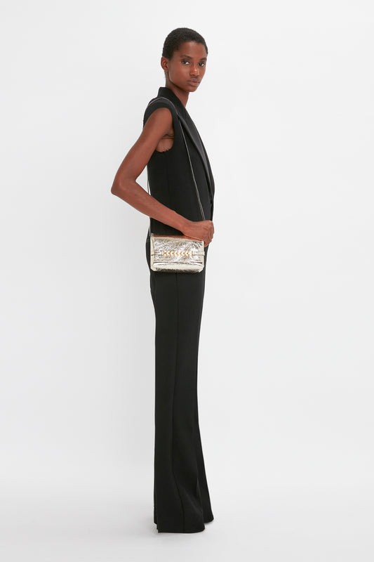 A woman in a black sleeveless jumpsuit stands sideways, looking at the camera, holding a Victoria Beckham Mini Chain Pouch With Long Strap In Gold Leather.