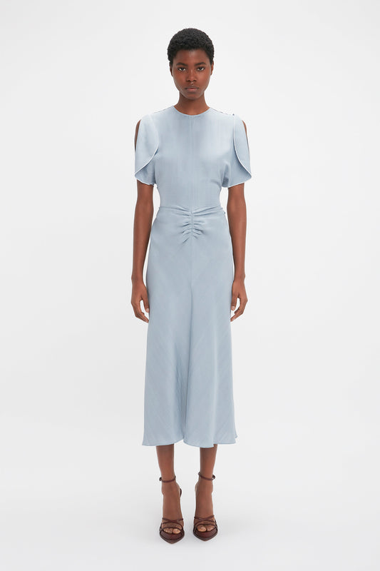 A woman stands directly facing the camera, wearing a pale blue Exclusive Gathered Waist Midi Dress In Pebble by Victoria Beckham with cut-out shoulders and brown heeled sandals.