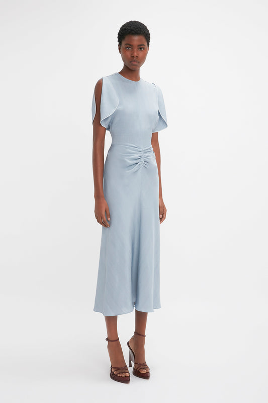 A woman stands against a white background wearing a light blue, knee-length Exclusive Gathered Waist Midi Dress In Pebble with ruffle sleeves and paired with brown heels by Victoria Beckham.
